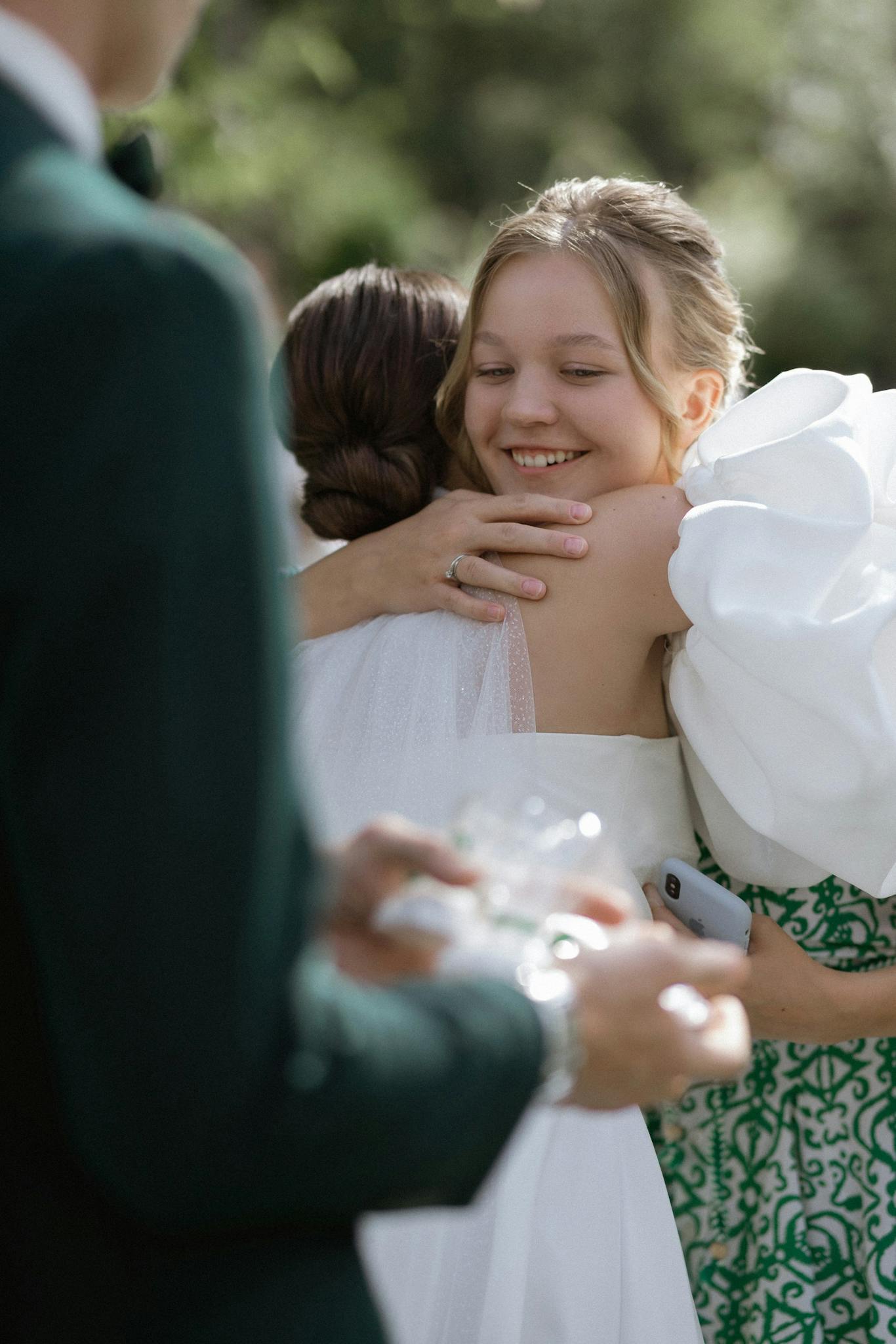 Smiling bridesmaid hugging bride at outdoor wedding ceremony, filled with joy and warmth.